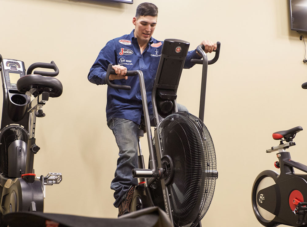 Jess Pope lleva una camisa azul abotonada y pantalones vaqueros azules, y monta una bicicleta fija para calentar antes de competir en un rodeo.